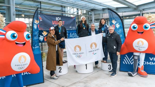 les deux mascottes des JO Paris 2024 encadrent 3 femmes médaillées sur un podium les membres de Paris 2024 tiennent devant elles un drapeau Paris 2024 
